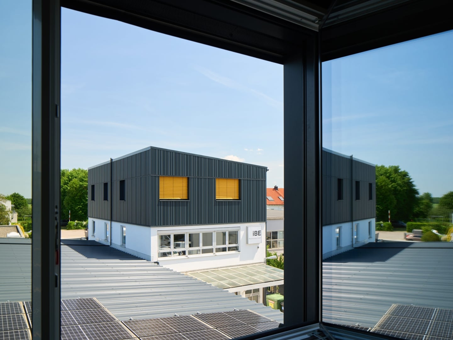 Blick durch ein offenes Fenster auf ein benachbartes Firmengebäude mit grauer Fassade und gelben Raffstores