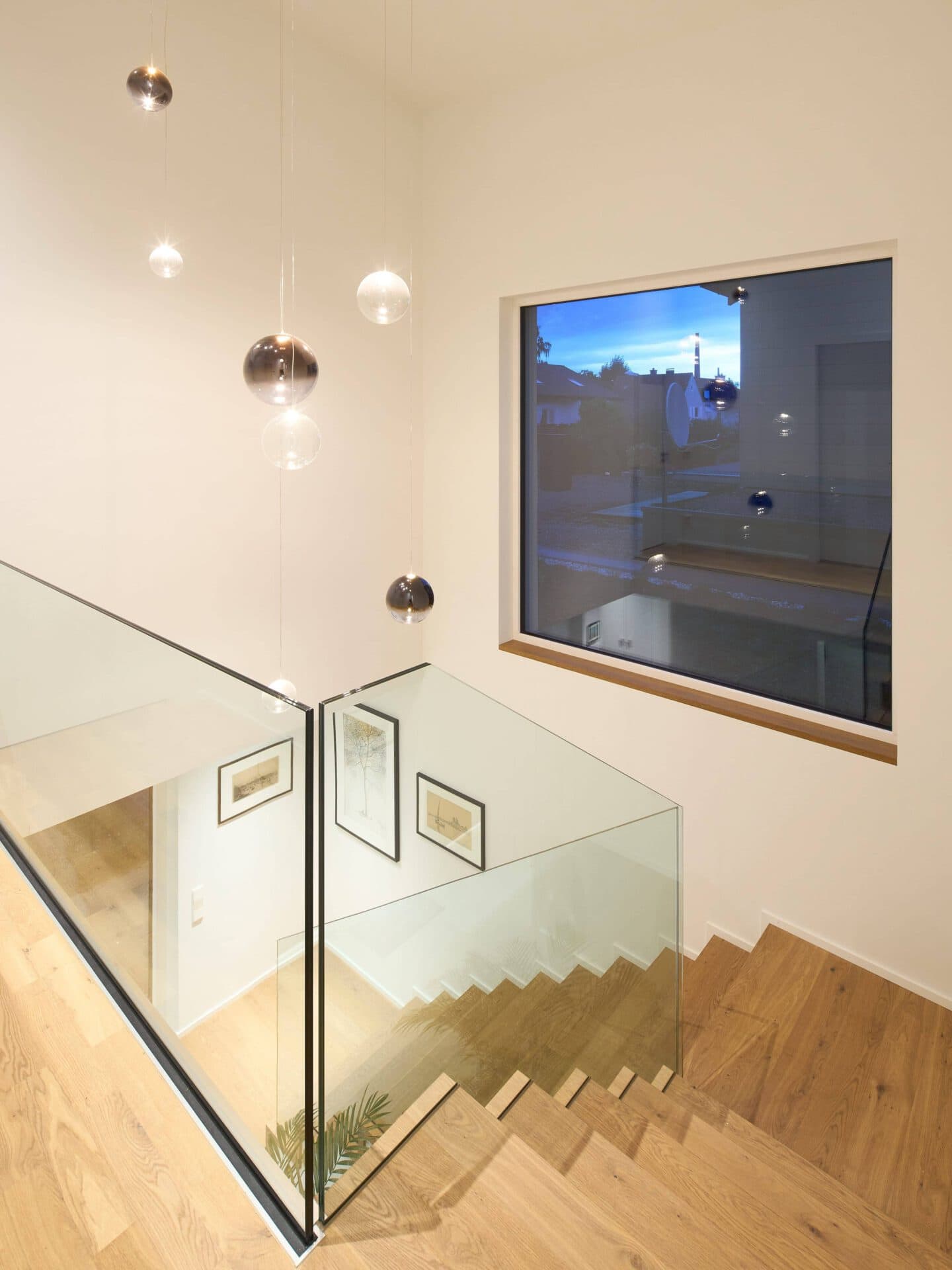Treppenhaus eines modernen Einfamilienhauses mit Holzstufen, Glasgeländer und großem Fenster.
